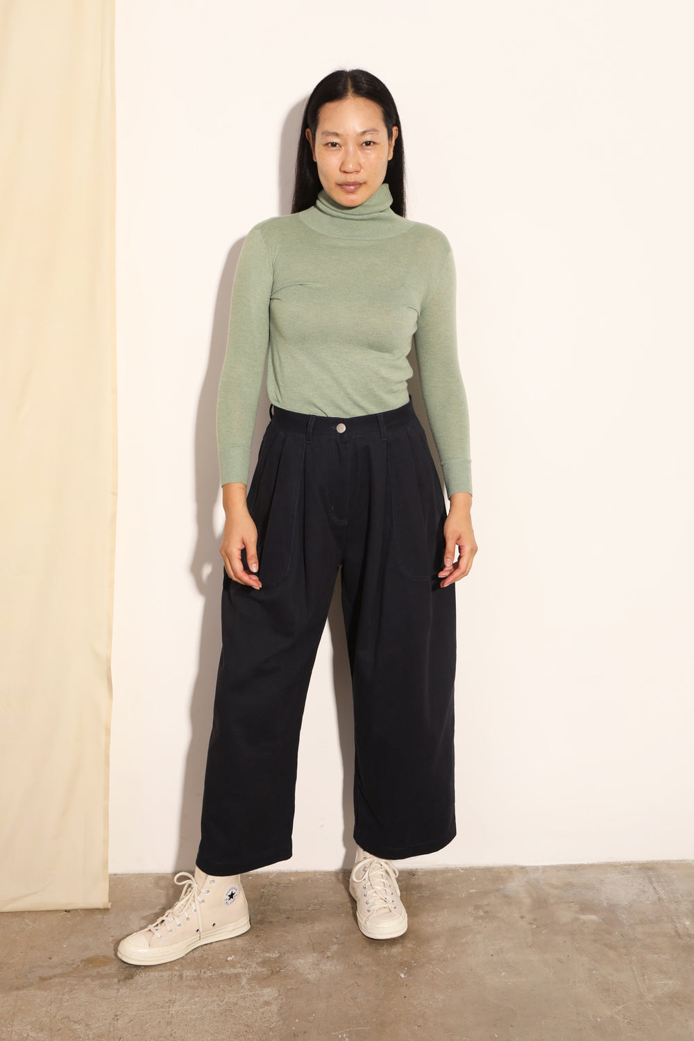 Person wearing a green turtleneck and black pants against a white wall.