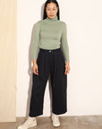 Person wearing a green turtleneck and black pants against a white wall.